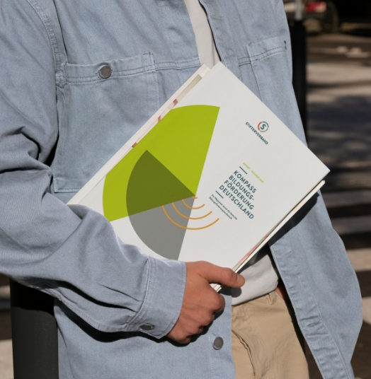 Report Design Kompass Bildungsförderung: Der Report wird als gedruckte Veröffentlichung von einer Person unter dem Arm gehalten. Das Cover des Reports vom Stifterverband ist sichtbar.