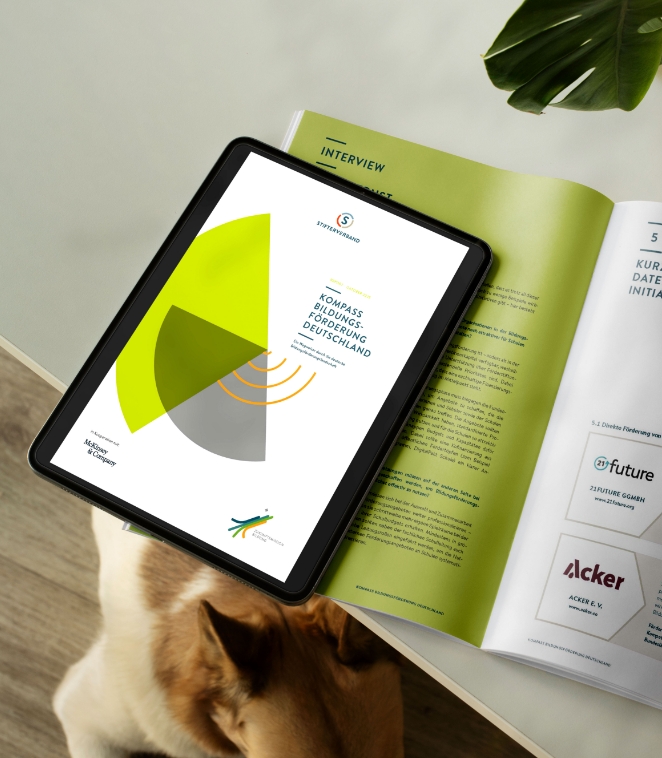 Report Design Kompass Bildungsförderung: Der Report liegt aufgeschlagen auf einem Tisch. Die Doppelseite zeigt links ein Interview und rechts Steckbriefe. Daneben liegt ein Tablet, welches den Report als digitale Veröffentlichung zeigt.